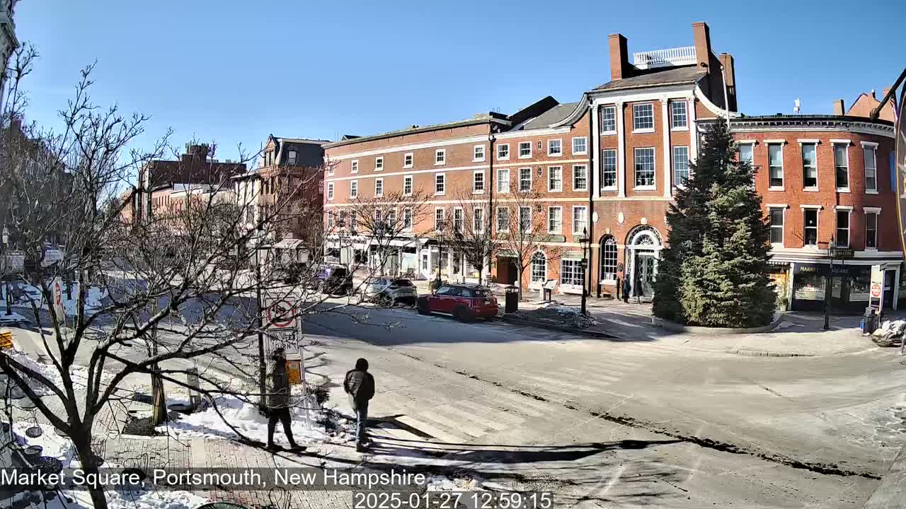 A sunny day reveals a town square with several multi-story brick buildings, a few cars, pedestrians, and patches of snow on the ground.