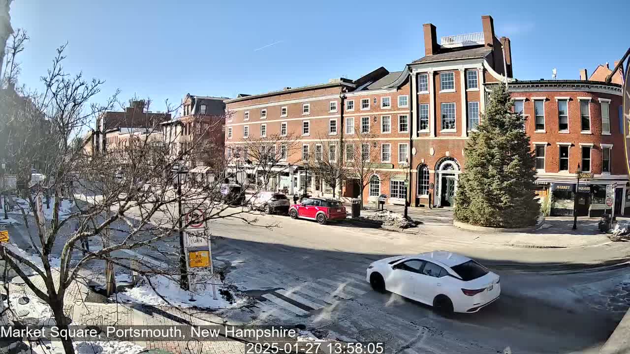 A sunny day in a city square shows several brick buildings, bare trees, some cars, and patches of snow and ice on the ground.