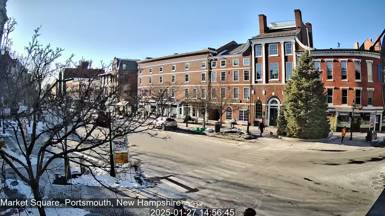 A sunny day in a city square with brick buildings, some patches of snow and ice on the ground, and a few pedestrians and cars.