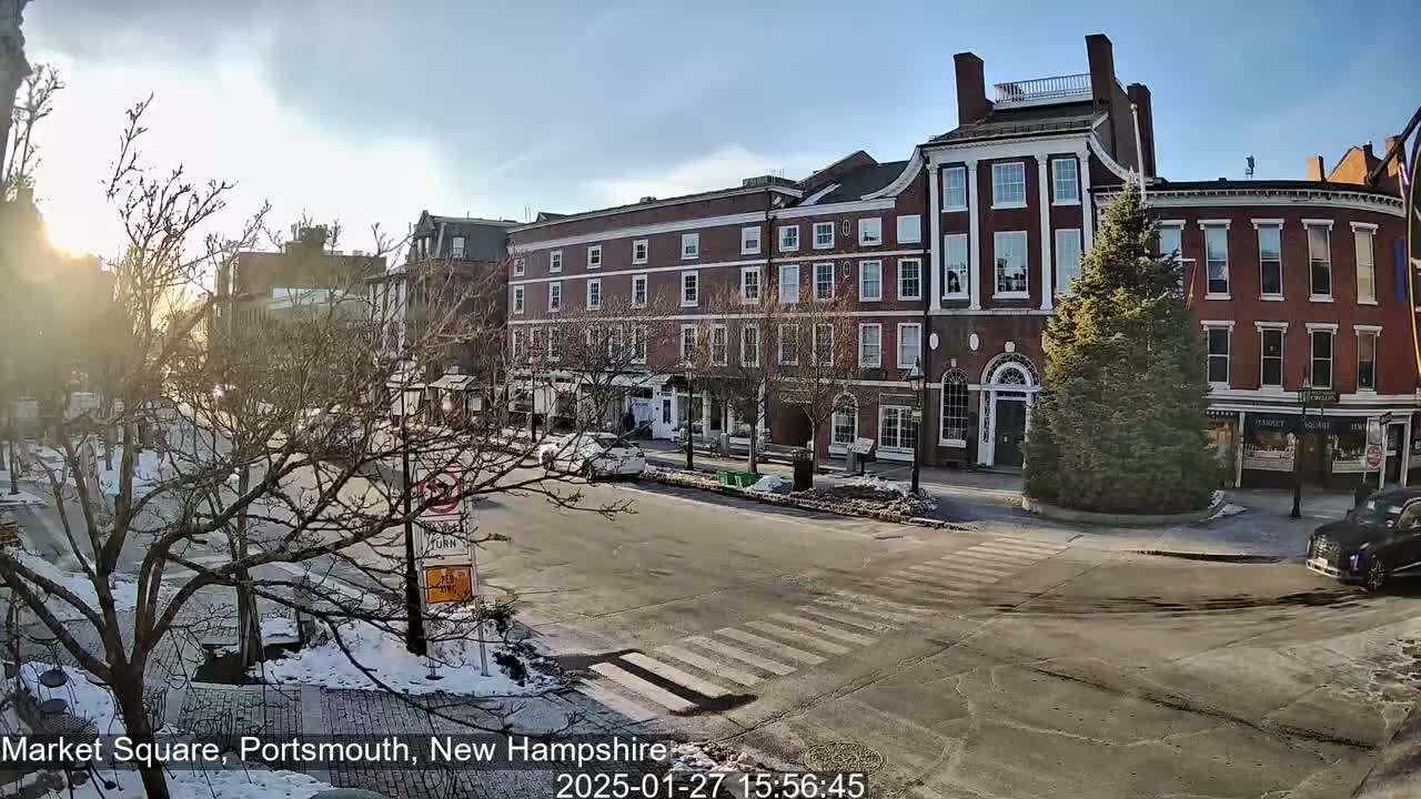 A sunny winter day in a town square shows several brick buildings, bare trees, a few cars, and some snow on the ground.