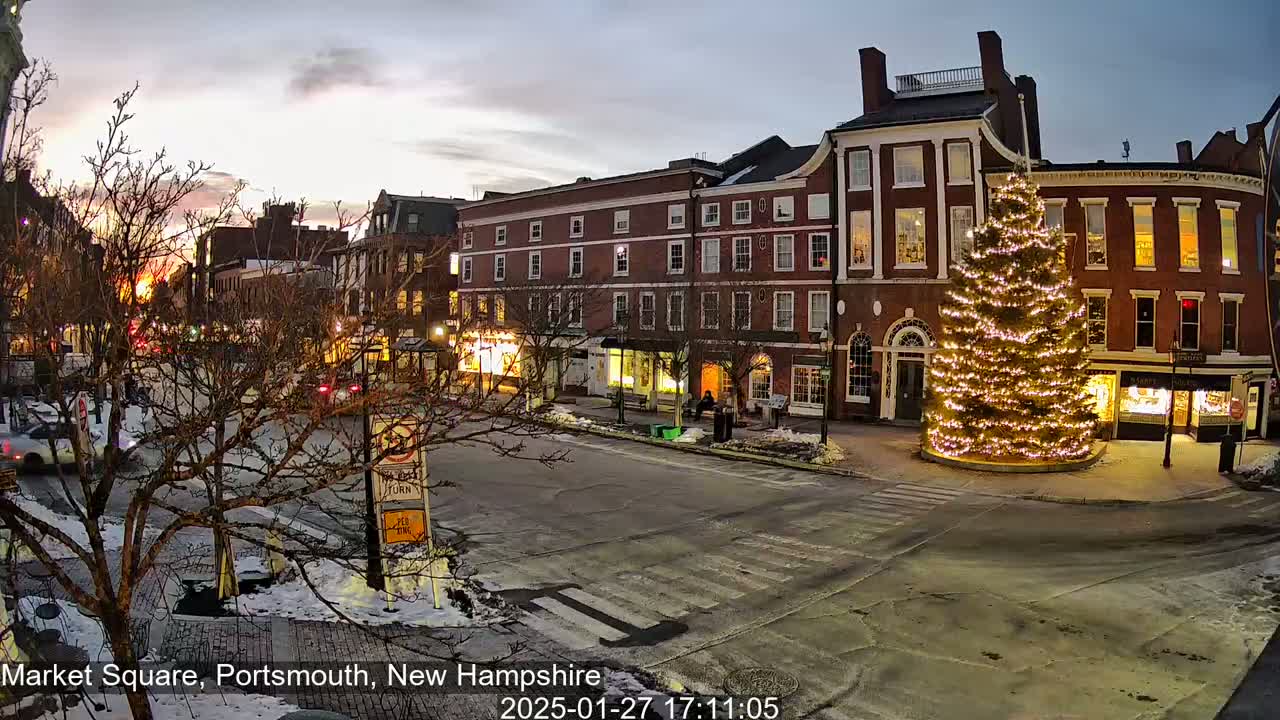 A large, illuminated Christmas tree stands in a snow-dusted town square at twilight, surrounded by brick buildings and sparsely-trafficked streets.
