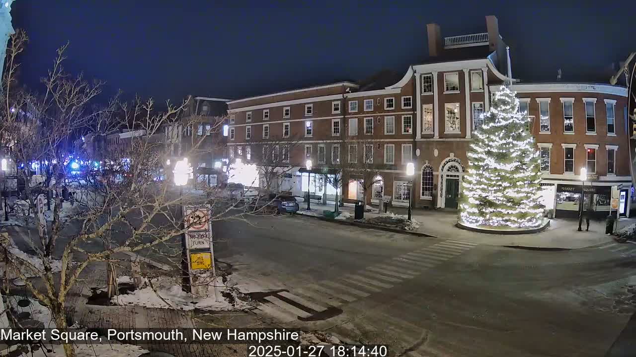 A snow-covered town square at night, illuminated by streetlights, features a large, decorated Christmas tree in the center and several brick buildings lining the street.