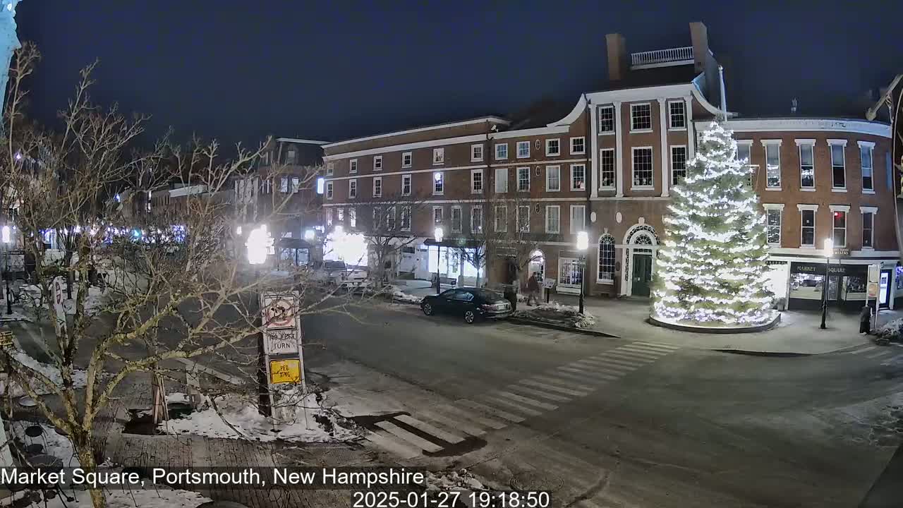 A snow-dusted market square at night, illuminated by streetlights, features a large, brightly lit Christmas tree in the center, with brick buildings lining the square.