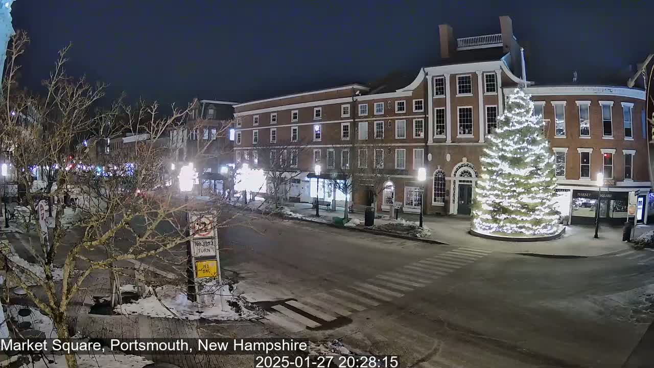 A snow-dusted night scene shows a town square with a large, illuminated Christmas tree in the center, surrounded by brick buildings and sparsely-traveled streets.