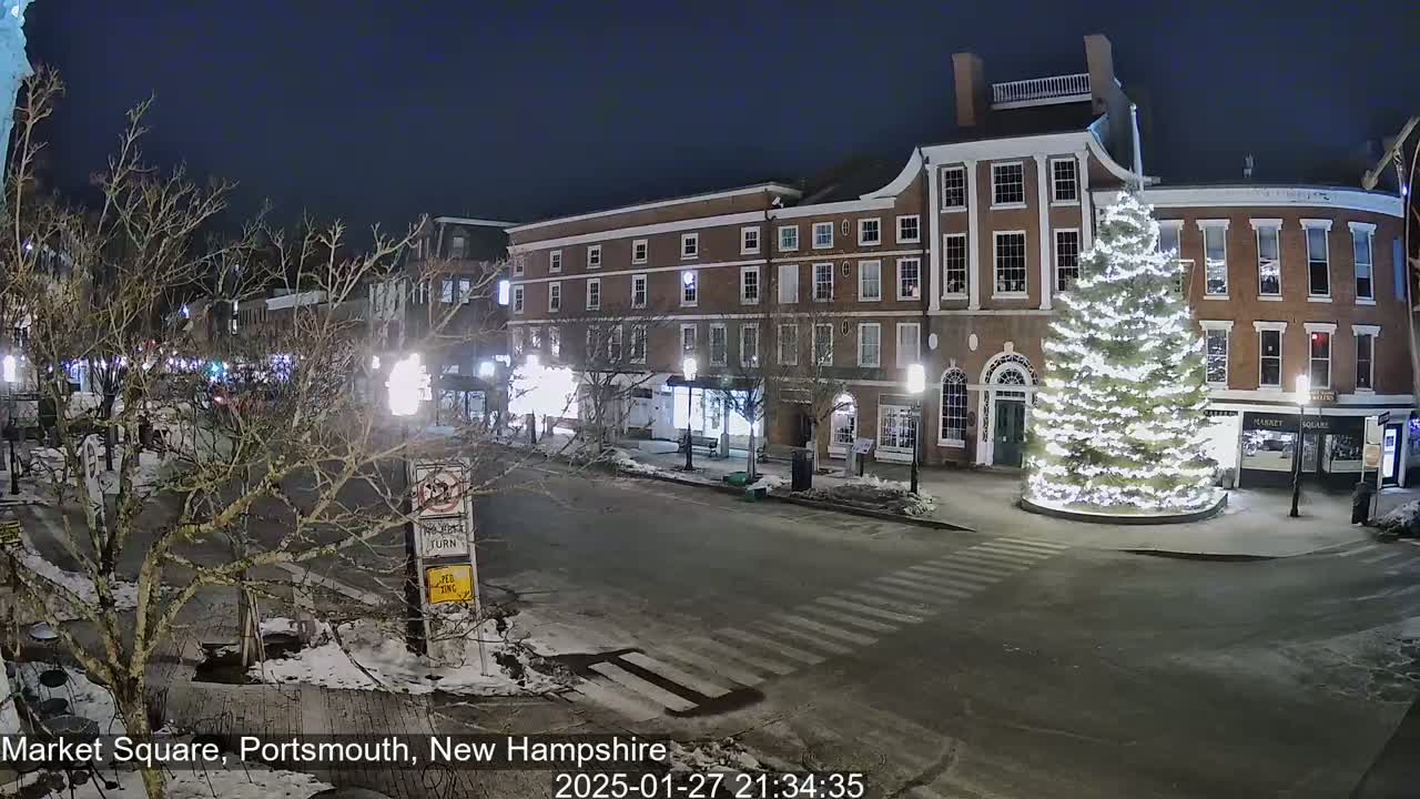 A snow-covered nighttime street scene shows a large illuminated Christmas tree in a town square, surrounded by brick buildings.