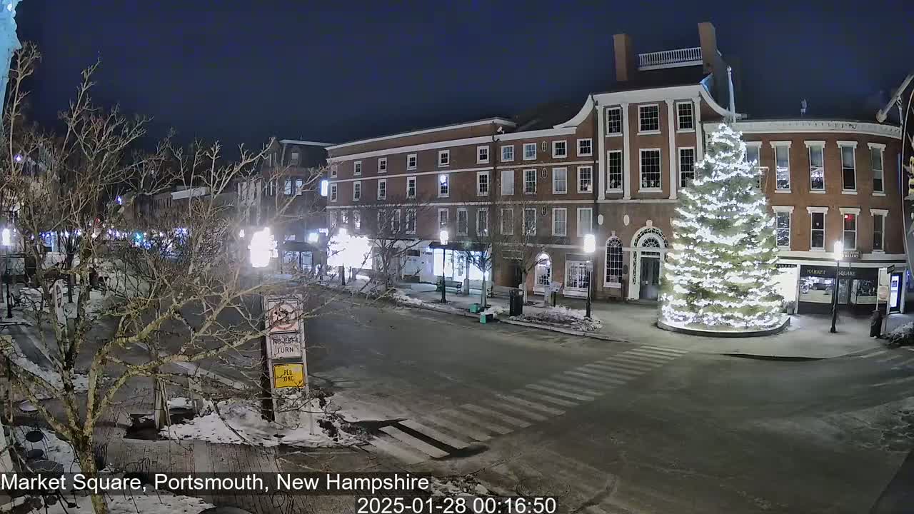 A snow-dusted, nighttime scene shows a town square with a large, illuminated Christmas tree in front of brick buildings.