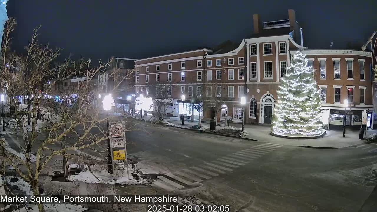 A snow-covered Market Square at night, illuminated by streetlights, features a large, brightly lit Christmas tree in the center, surrounded by brick buildings.