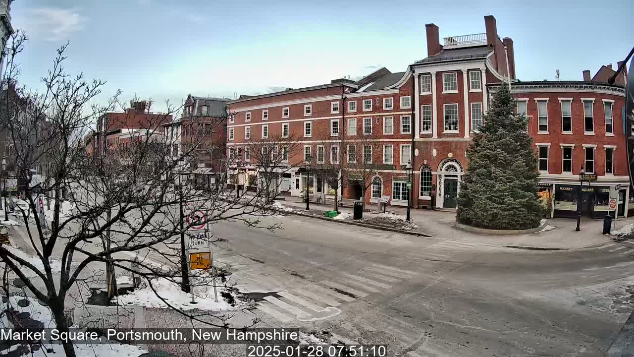 A mostly empty town square with snow-covered sidewalks and roads, featuring several brick buildings under a partly cloudy sky.