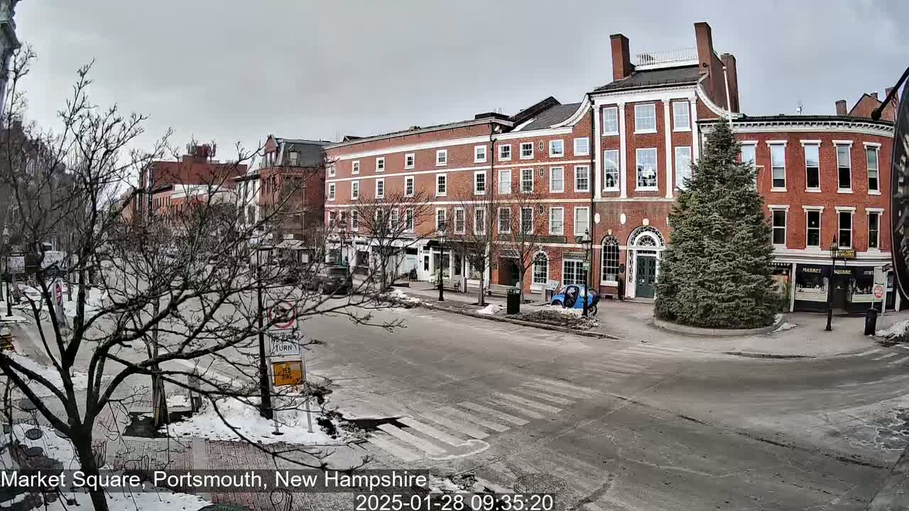 A mostly empty street scene with several brick buildings, bare trees, and a little snow on the ground under an overcast sky.