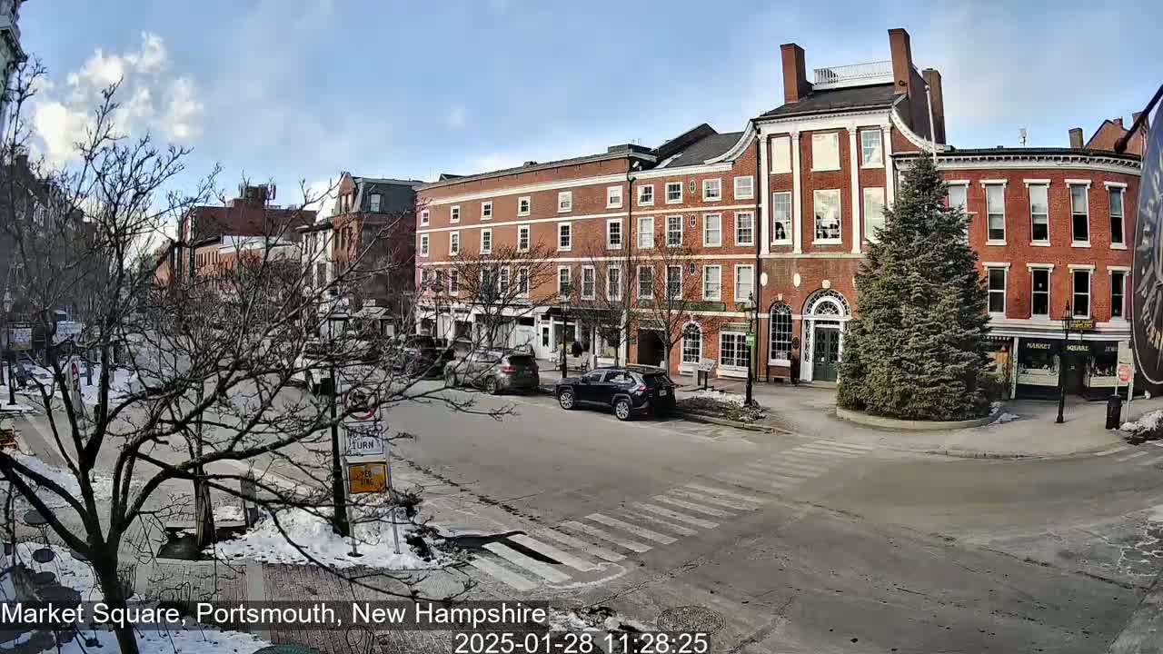A sunny winter day in a town square, showing brick buildings, parked cars, and a snow-covered street with a pedestrian crossing.