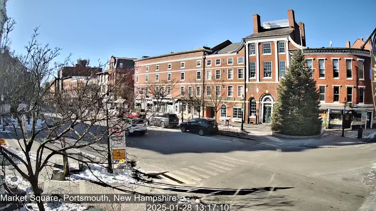 A sunny day in a city square shows several brick buildings, a few cars, a small evergreen tree, and patches of snow on the ground.