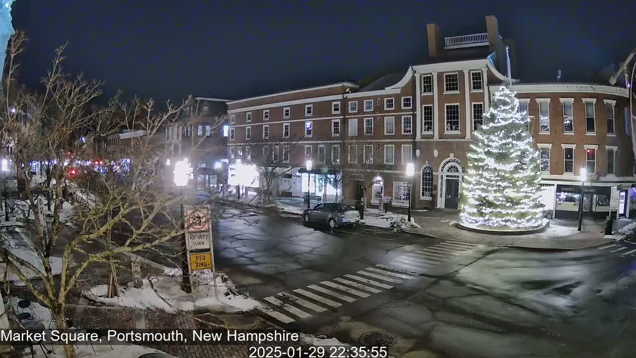 A snow-covered town square at night features a large illuminated Christmas tree in the center, surrounded by brick buildings and a few cars.