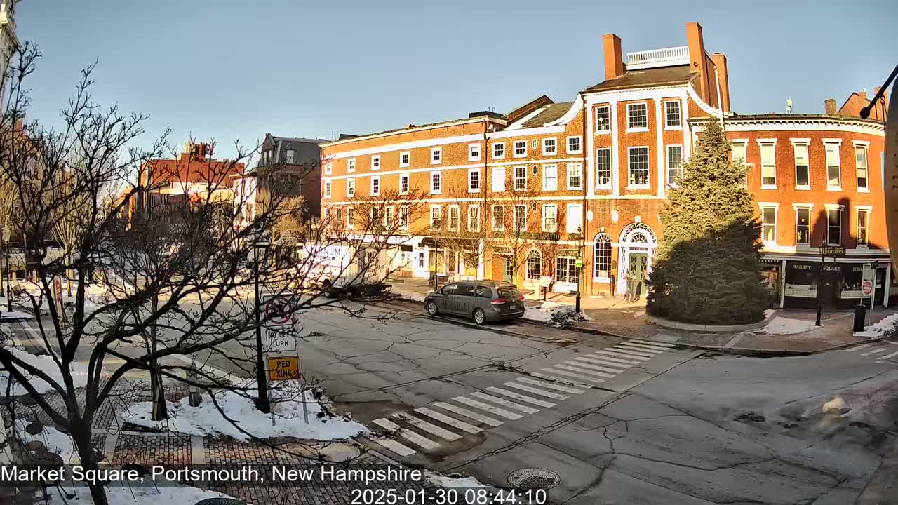 A sunny winter day shows a brick building and a street with a crosswalk, patchy snow on the ground, and bare trees.
