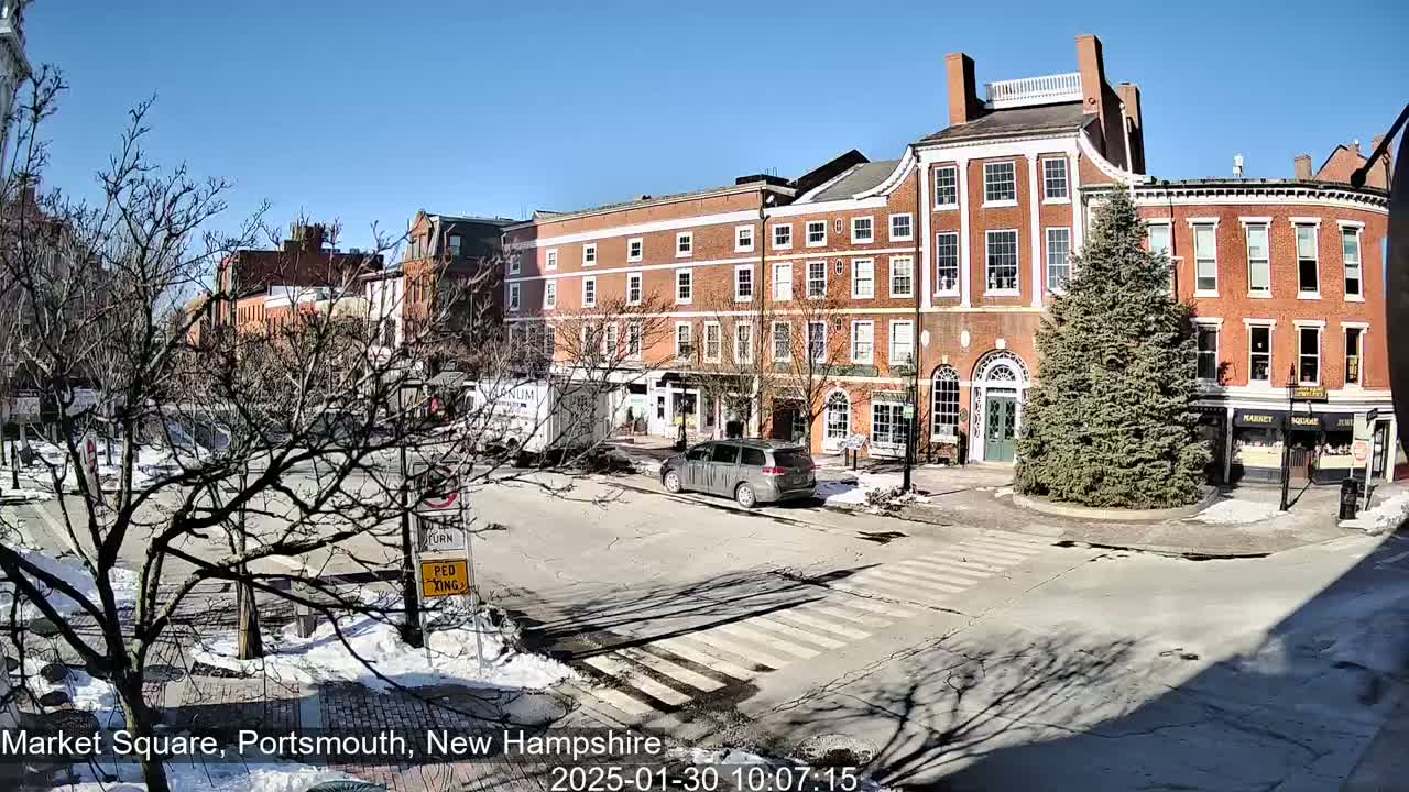 A sunny day reveals a city square with several brick buildings, a minivan, and patchy snow on the ground.
