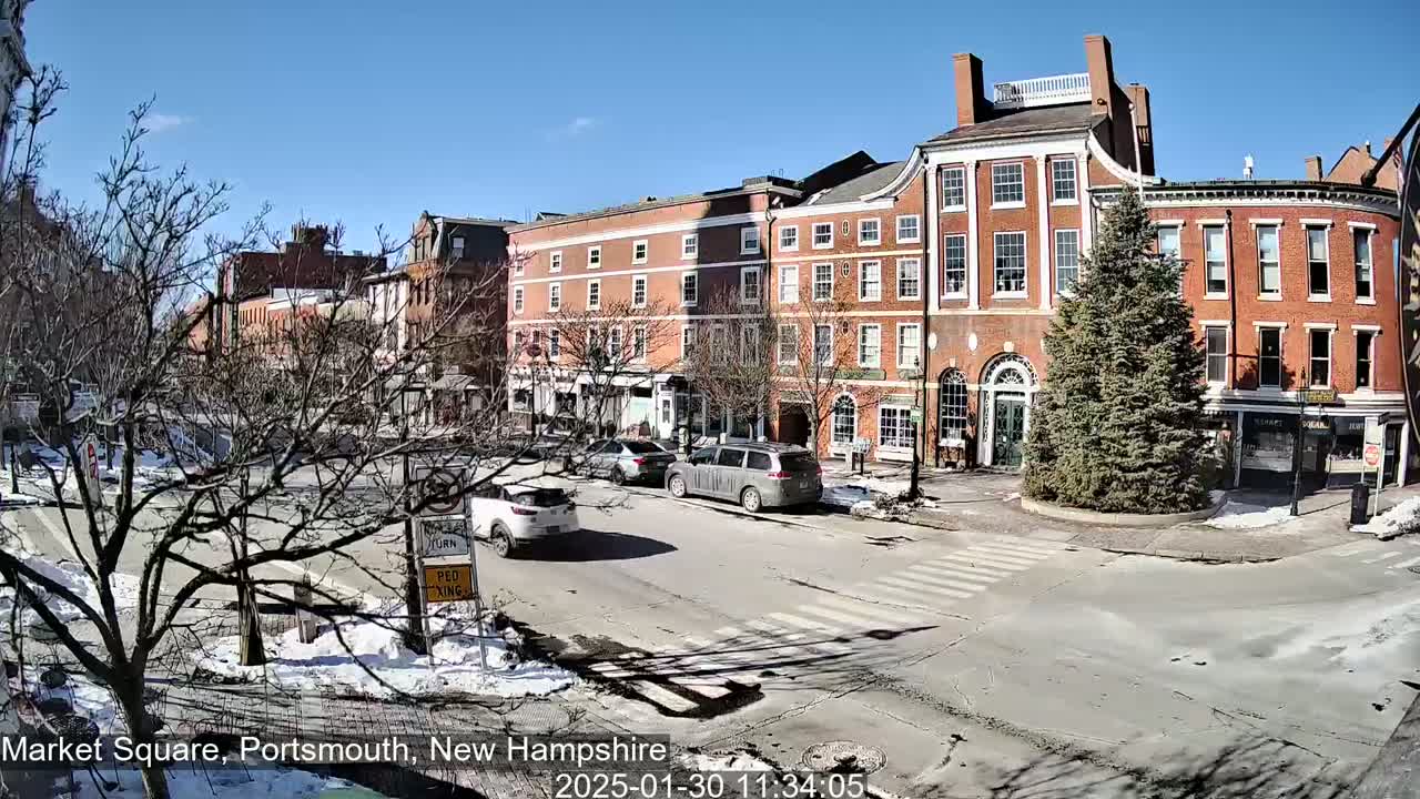 A sunny day in a city square with several brick buildings, a few cars, and patches of snow on the ground.