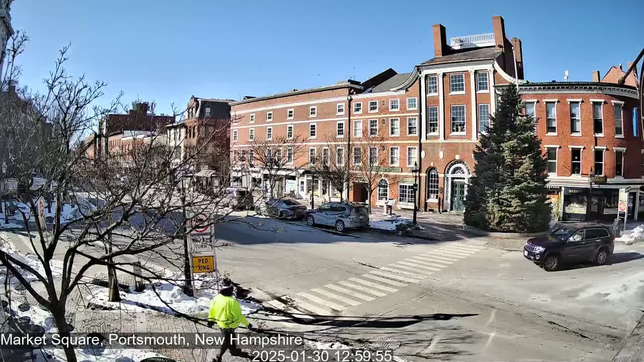A sunny day in a town square with several brick buildings, parked cars, a pedestrian, and patches of snow on the ground.