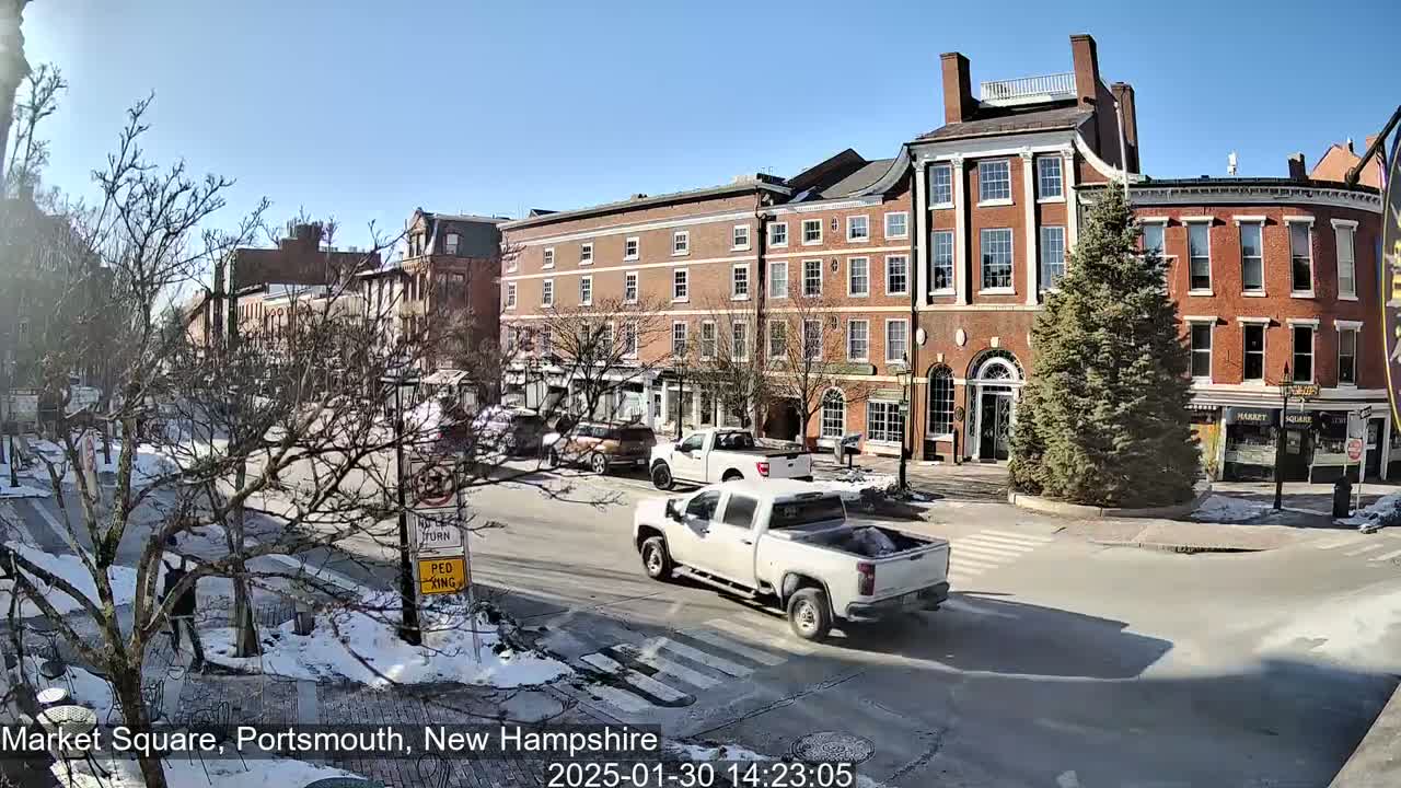 A sunny day in a town square shows several brick buildings, cars, and patches of snow on the ground.