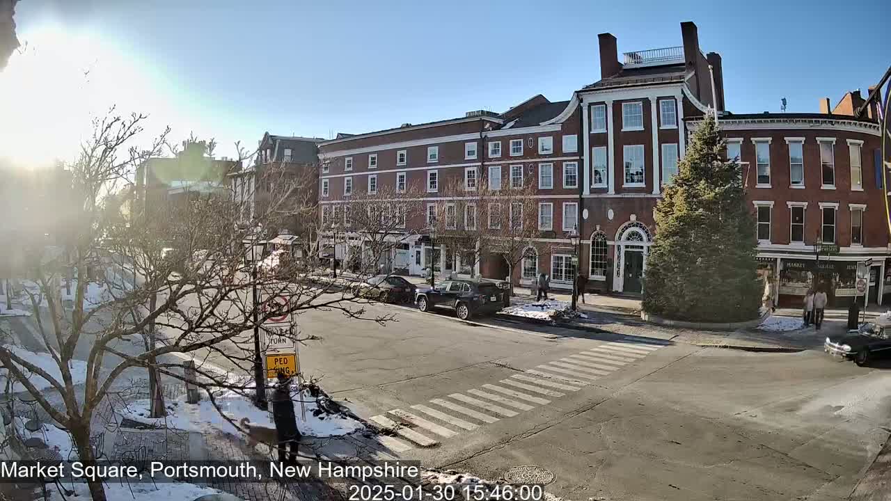 A sunny winter day in a town square shows several brick buildings, cars, pedestrians, and bare trees with some snow on the ground.