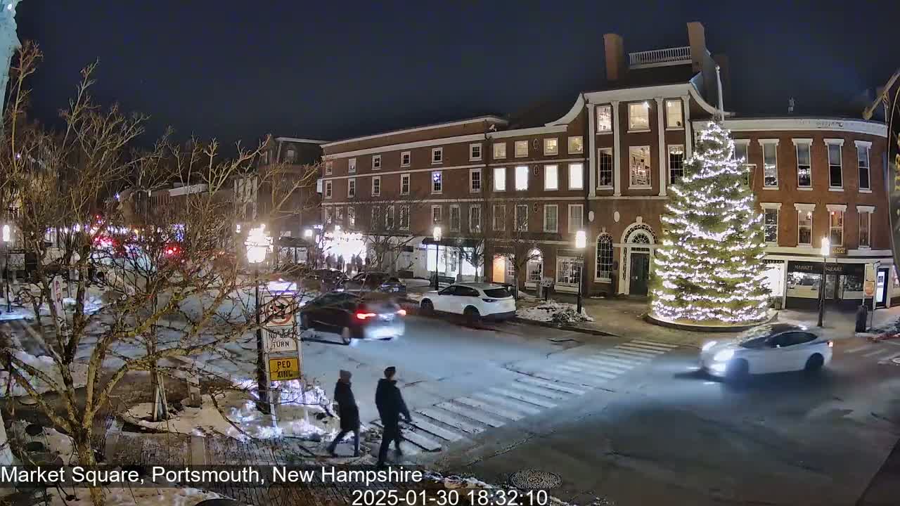 A snow-dusted night scene shows a brightly lit Christmas tree in a town square with cars driving by and two pedestrians crossing the street in front of brick buildings.