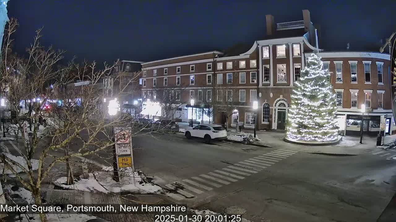 A snow-dusted town square at night, illuminated by streetlights, features a large, decorated Christmas tree in the center and several brick buildings with lit windows.