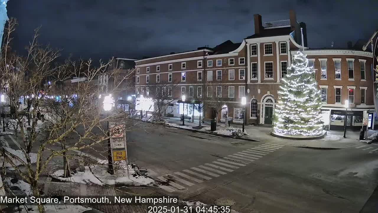 A snow-covered Market Square at night, featuring a large, illuminated Christmas tree in the center and brick buildings lining the street.