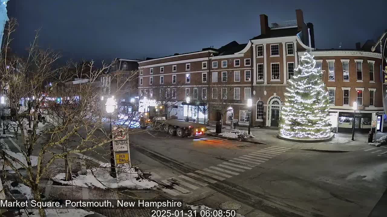 A snow-covered town square at night features a large, illuminated Christmas tree in the center, with brick buildings lining the street and a truck driving past.