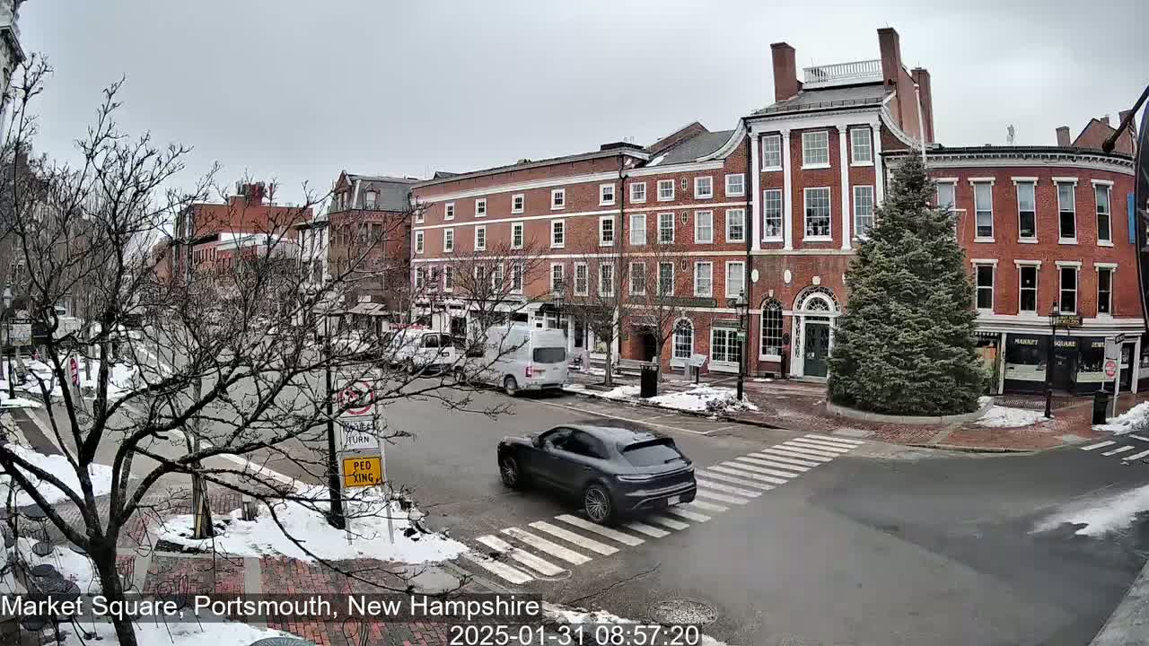 A dark-grey SUV crosses a pedestrian crossing in a snow-dusted town square, where several brick buildings stand under an overcast sky.