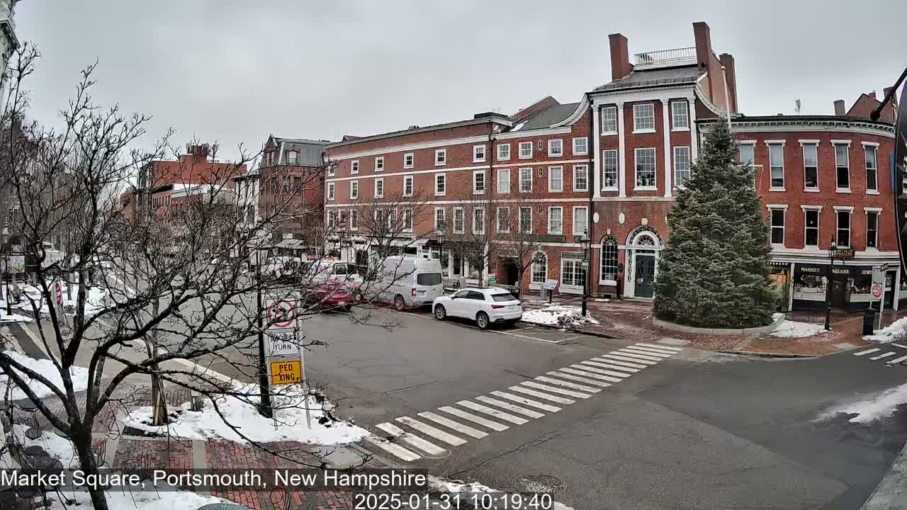 A street scene shows several brick buildings under an overcast sky with patches of snow on the ground and parked cars.