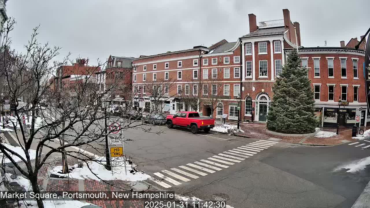 A red pickup truck is parked near a crosswalk in a snow-dusted town square with several brick buildings under an overcast sky.