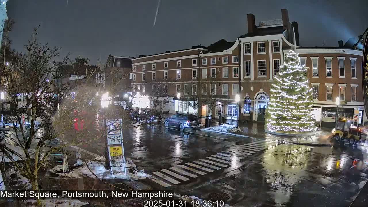 A wet, nighttime city square features a large illuminated Christmas tree in the center, surrounded by brick buildings, cars, and a street sweeper.