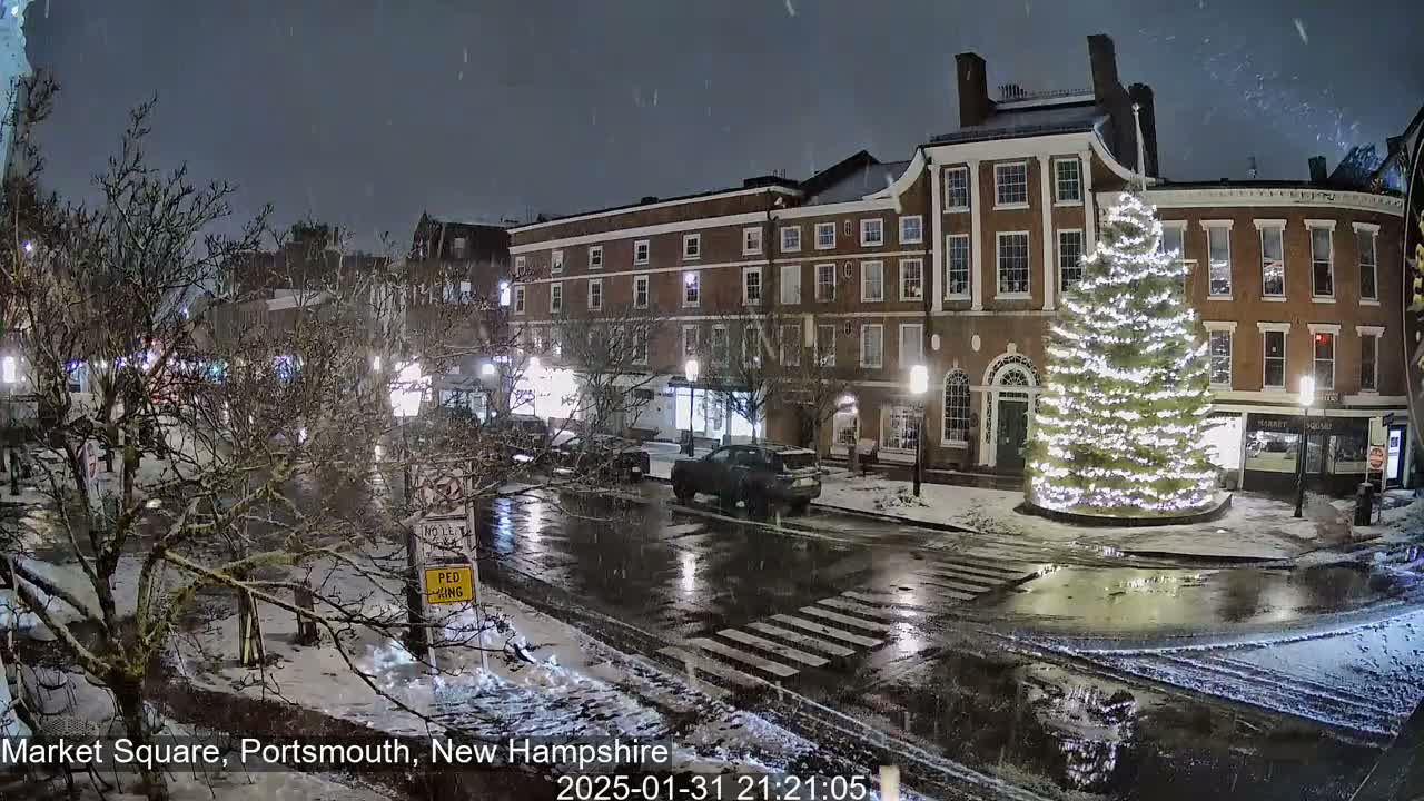 A snow-covered town square at night features a large illuminated Christmas tree in the center, with brick buildings lining the street and cars parked along the curb.