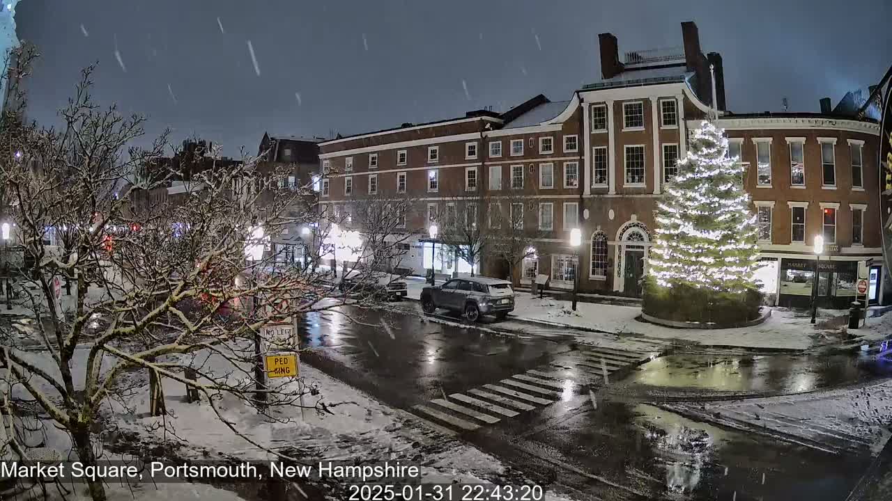 A snow-covered town square at night, featuring a large illuminated Christmas tree in front of brick buildings, with light snowfall continuing.
