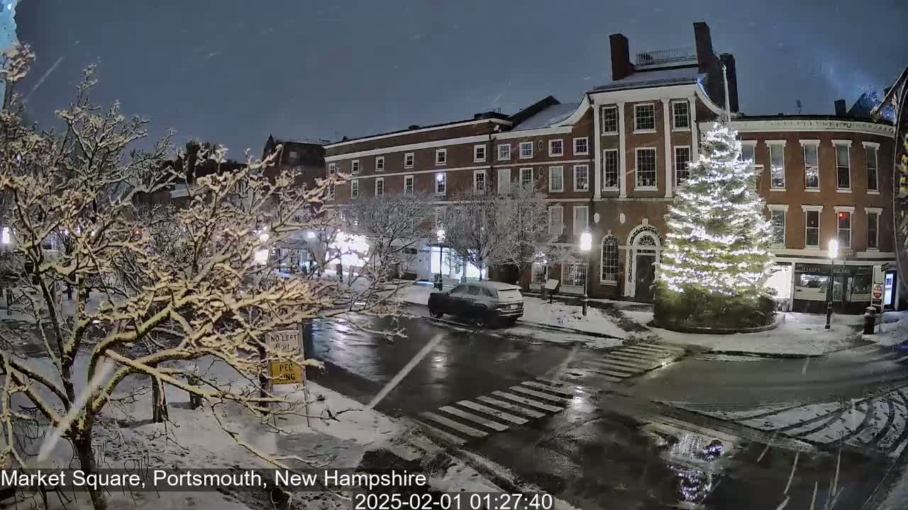 Snow falls at night on a town square with snow-covered trees, a decorated Christmas tree, and brick buildings.