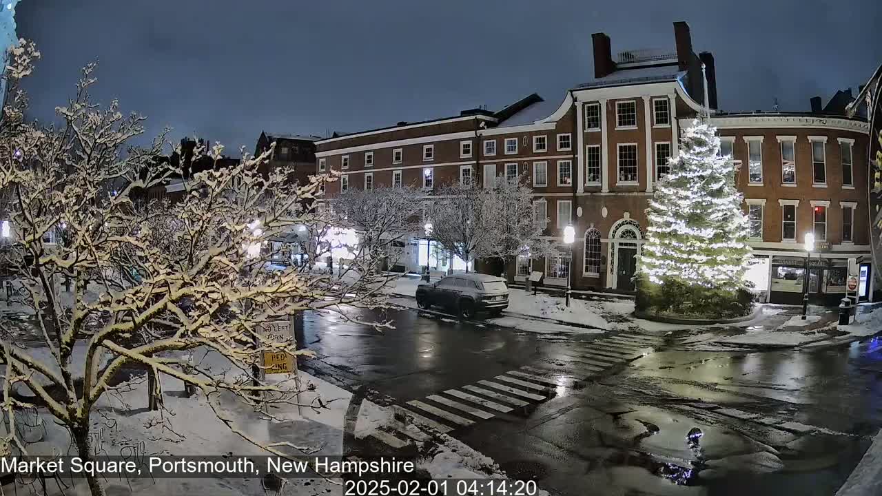 A snow-covered town square at night, featuring a large illuminated Christmas tree and a brick building, with wet roads and a vehicle parked nearby.