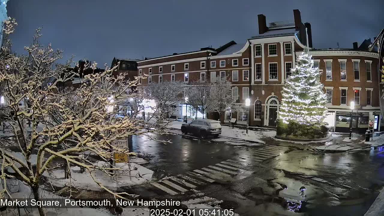 A snow-covered town square at night features a large illuminated Christmas tree, brick buildings, and a few cars, with wet patches on the street.