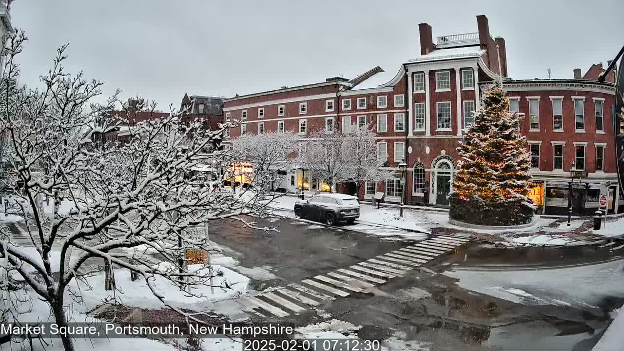 A snow-covered town square with brick buildings, a lit-up Christmas tree, and a vehicle parked nearby under a cloudy sky.