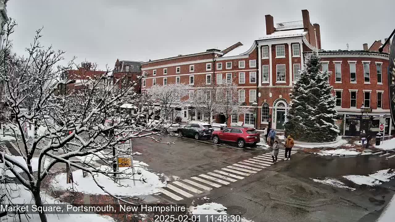 A snow-covered town square with brick buildings, parked cars, pedestrians crossing the street, and a large evergreen tree in the center.