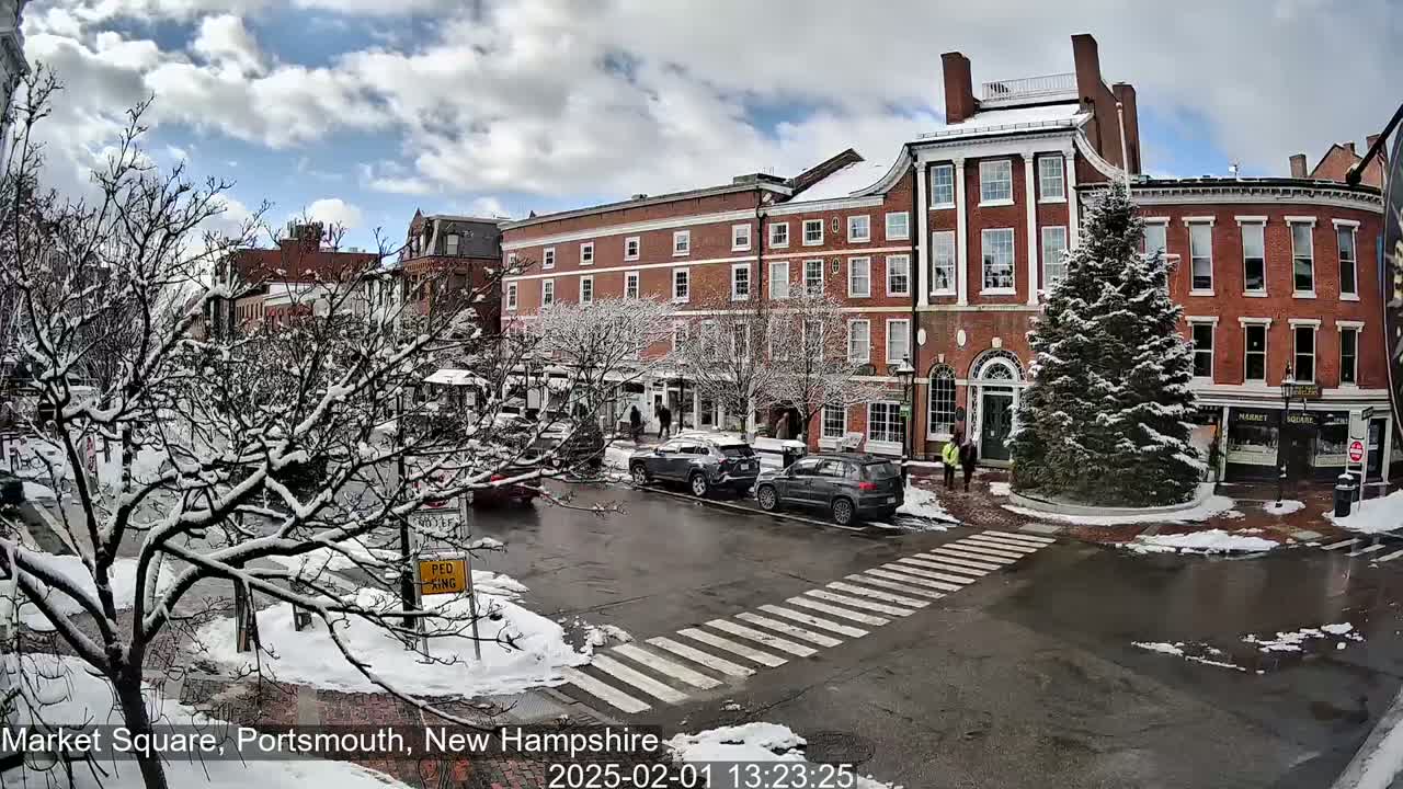 A snow-covered town square with brick buildings, parked cars, and a few pedestrians, under a partly cloudy sky.