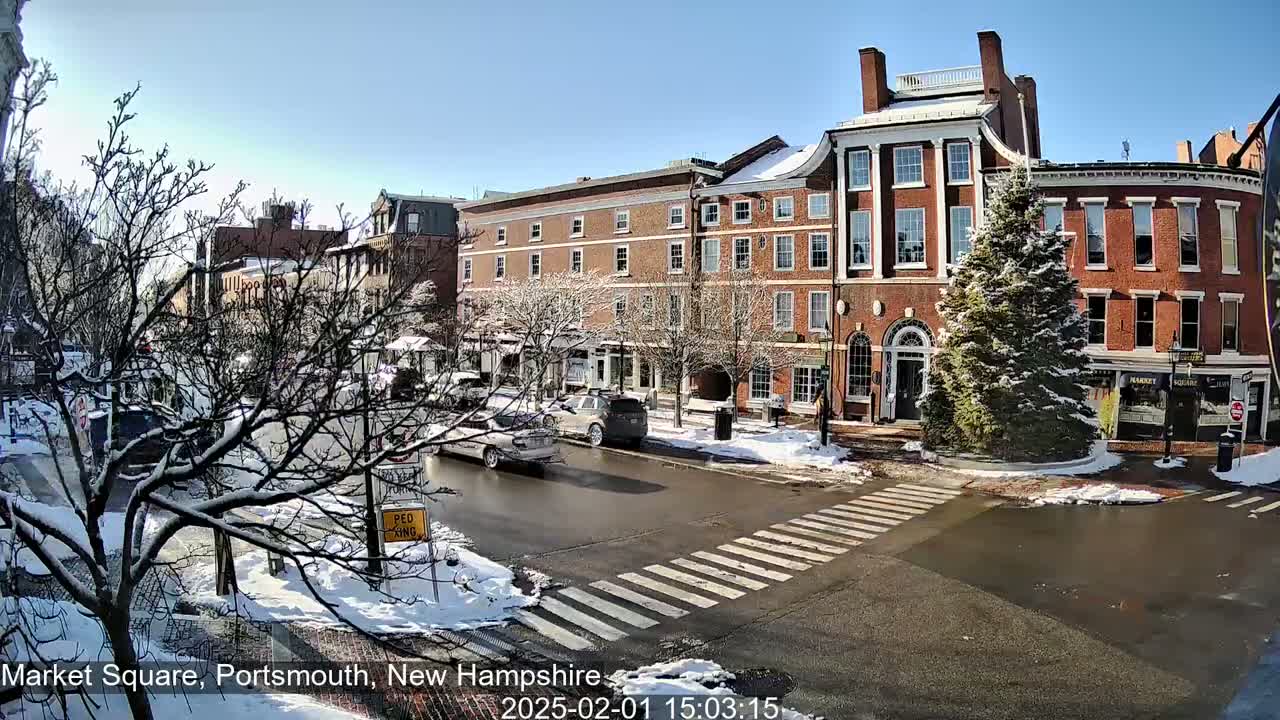 A snow-covered town square with brick buildings and several cars driving on a sunny day.