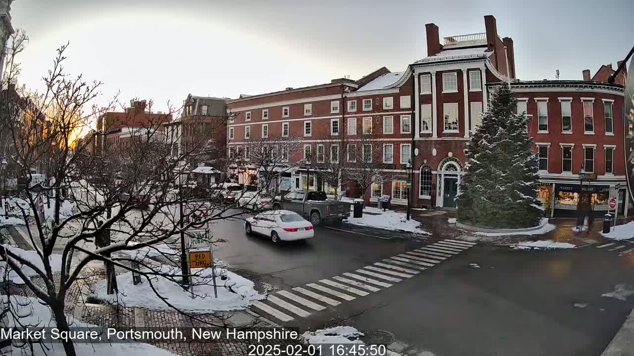 A snow-covered town square with brick buildings, cars driving, and a tree in the center, all under a dusky, post-sunset sky.