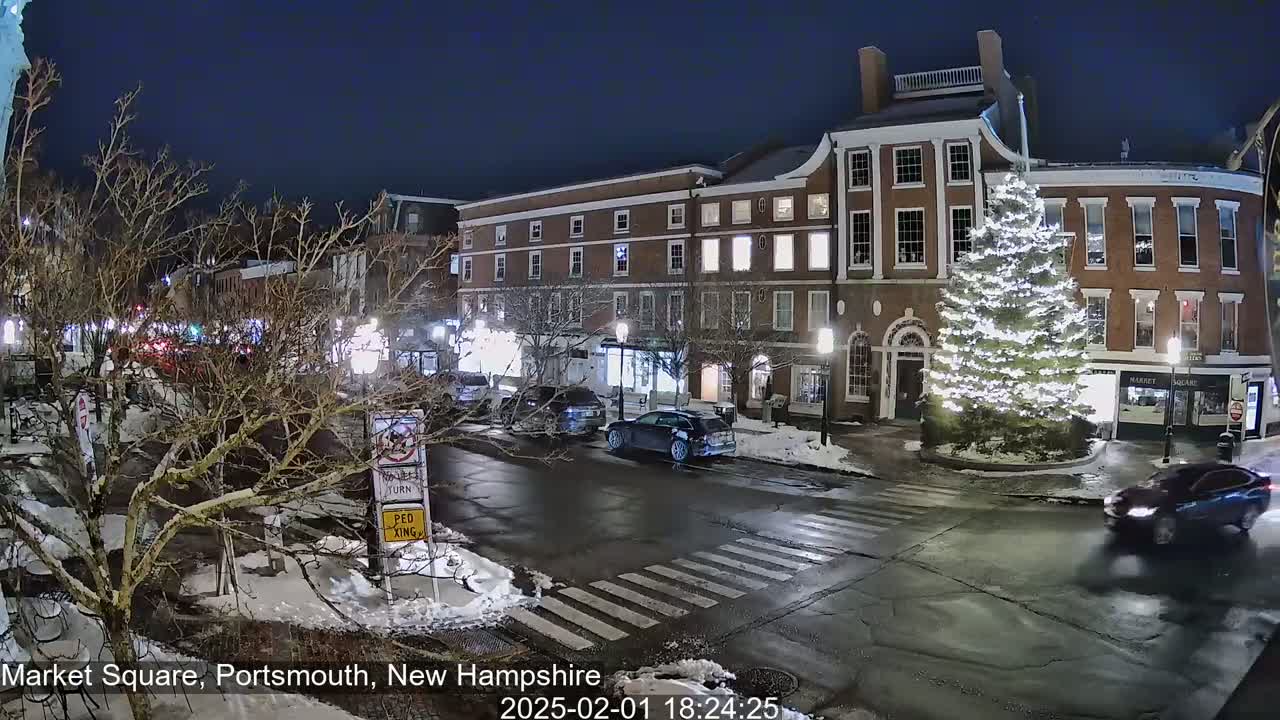 A nighttime scene of a snow-covered town square with a large, illuminated Christmas tree, several cars, and brick buildings.