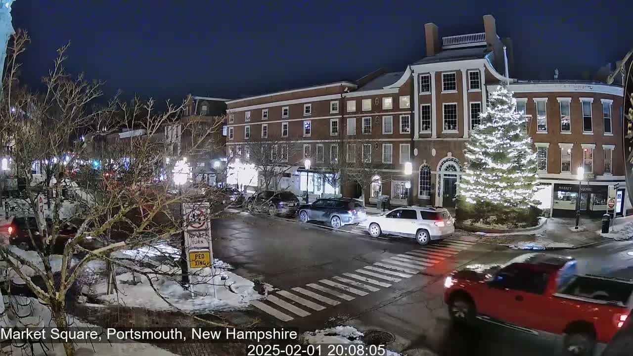 A snow-covered town square at night, featuring a large illuminated Christmas tree in front of brick buildings and several cars driving through a crosswalk.