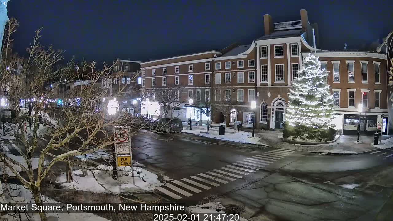 A snow-covered town square at night, illuminated by streetlights, features a large, lit Christmas tree in the center, with brick buildings lining the perimeter and sparsely parked cars.