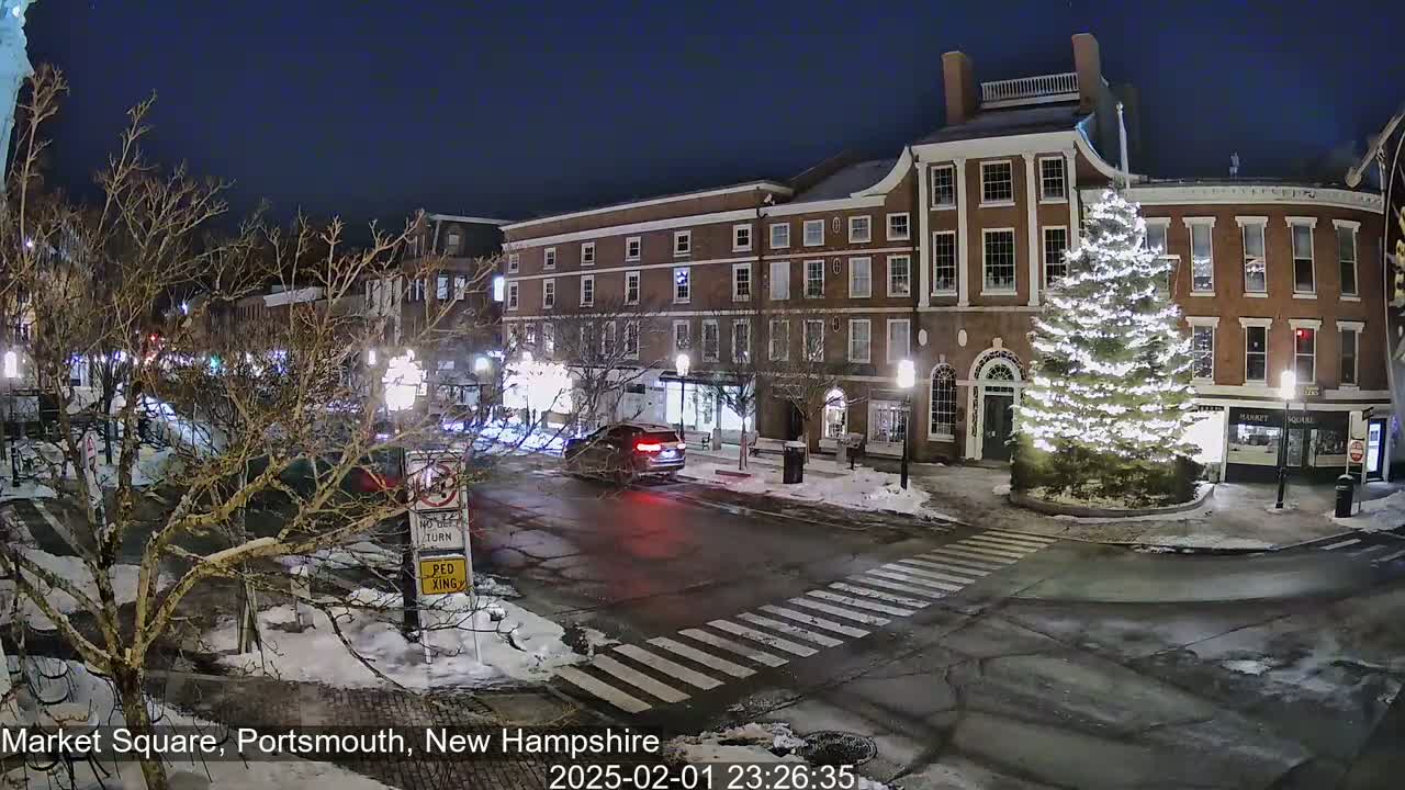 A snow-covered town square at night, illuminated by streetlights, features a large, illuminated Christmas tree in the center and brick buildings along the sides.