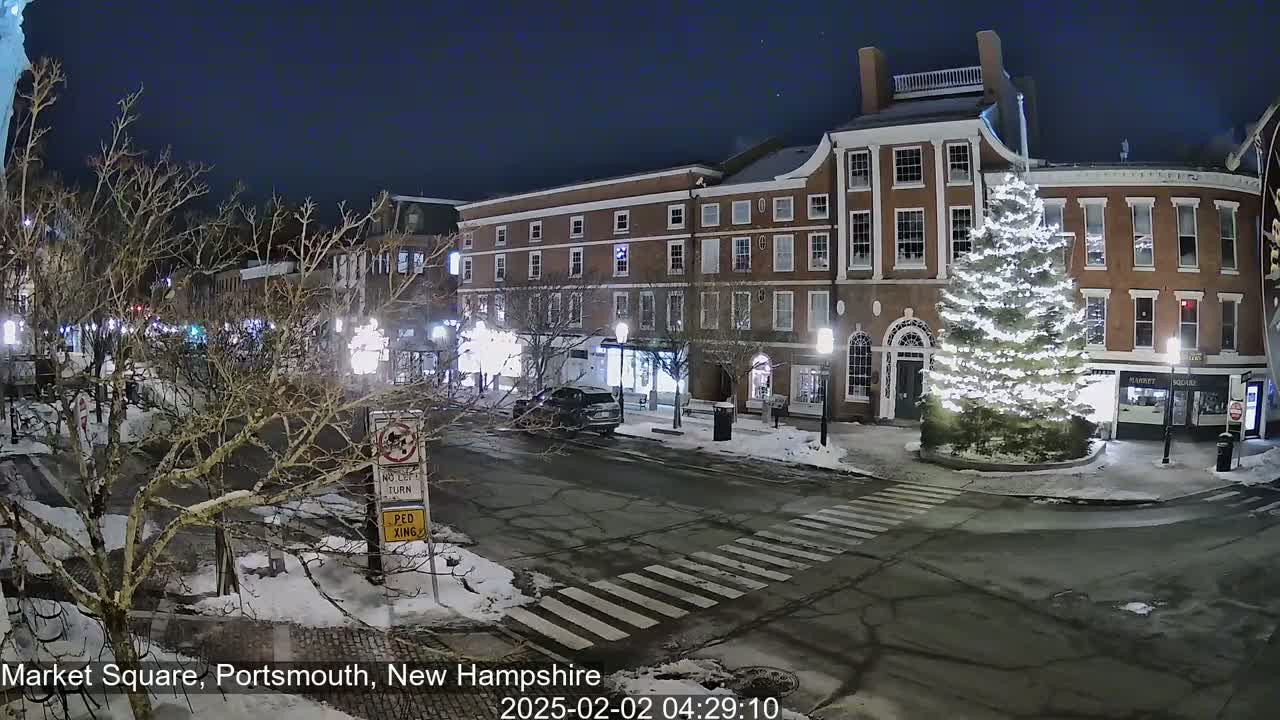 A snow-covered Market Square at night, illuminated by streetlights, features brick buildings, a large decorated Christmas tree, and a few cars.