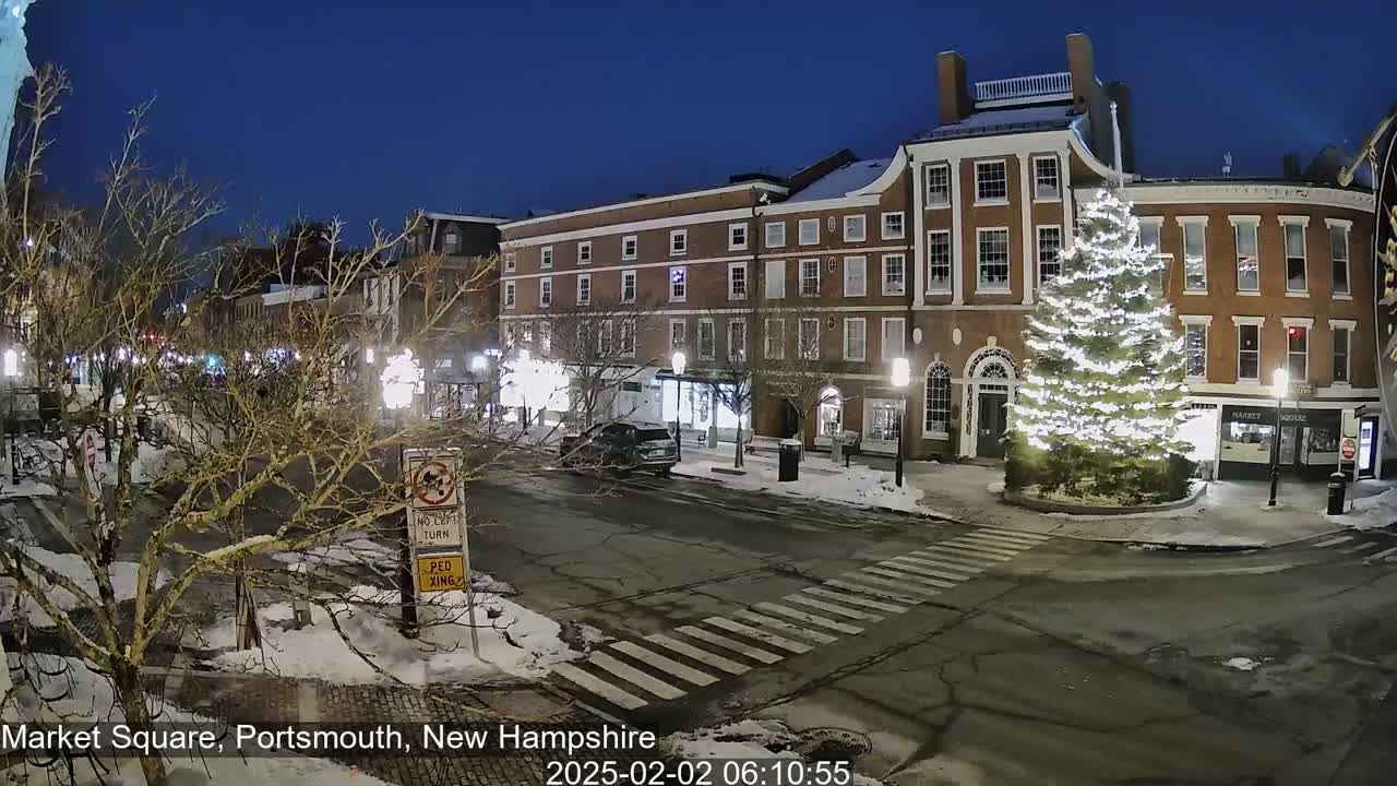A snow-covered town square at night, illuminated by streetlights, features a large decorated Christmas tree in front of brick buildings.