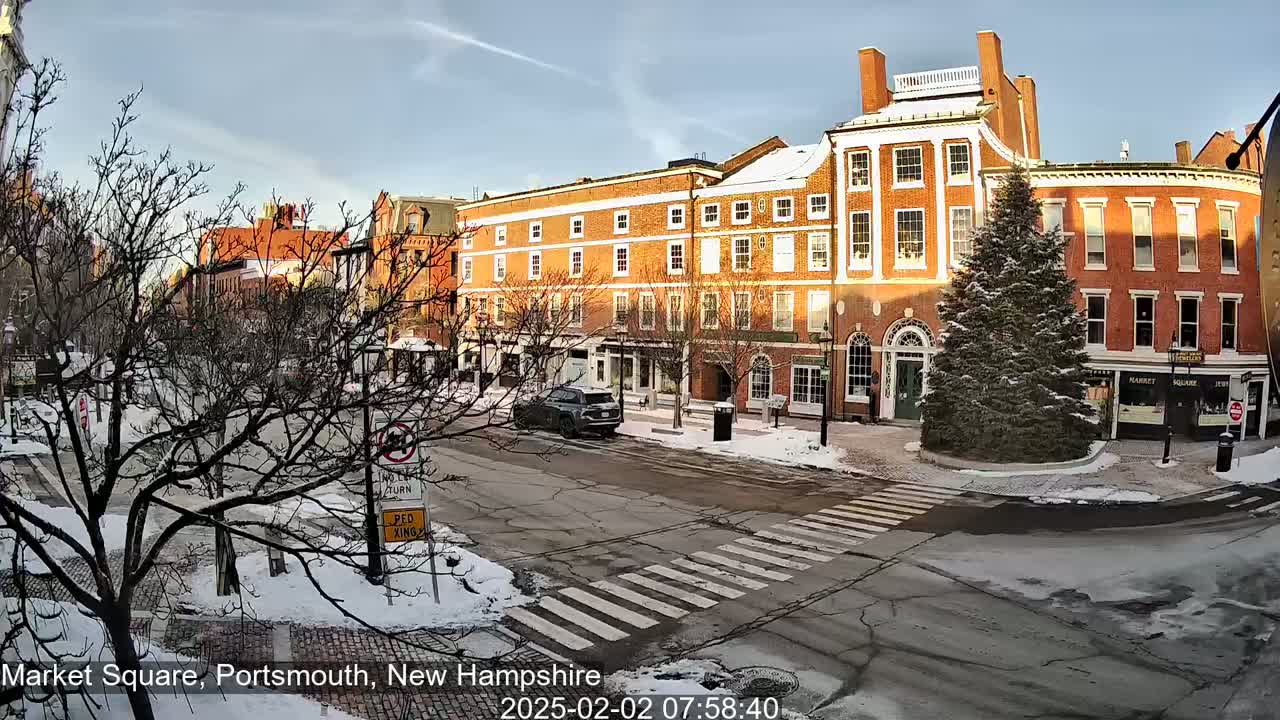 A snow-covered town square with brick buildings, a few cars, and a large evergreen tree in the center, under a mostly sunny sky.