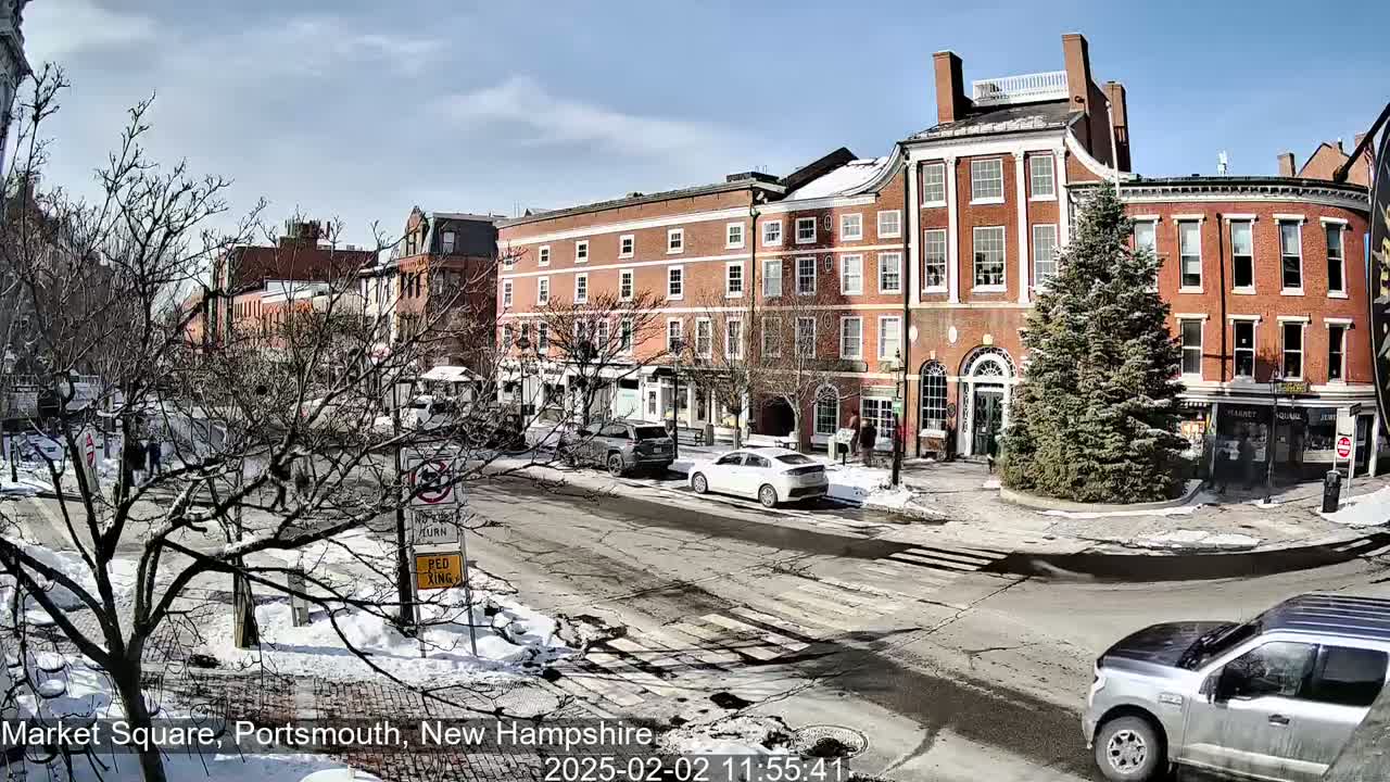 A snow-dusted street scene shows brick buildings, parked cars, and a few pedestrians under a sunny sky.