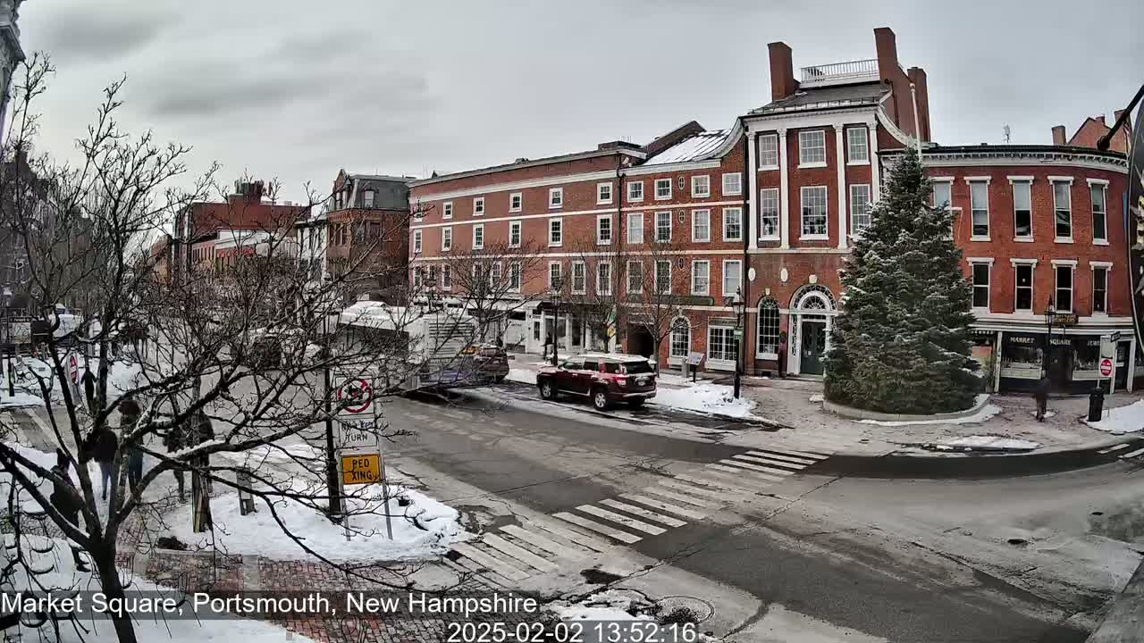 A snow-dusted town square with several brick buildings, a bus, a car, pedestrians, and a snow-covered evergreen tree in the center of the intersection.