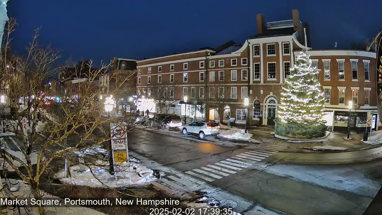 A snow-dusted Market Square at night shows a large, illuminated Christmas tree in the center of the street, in front of brick buildings with lit windows and cars driving by.